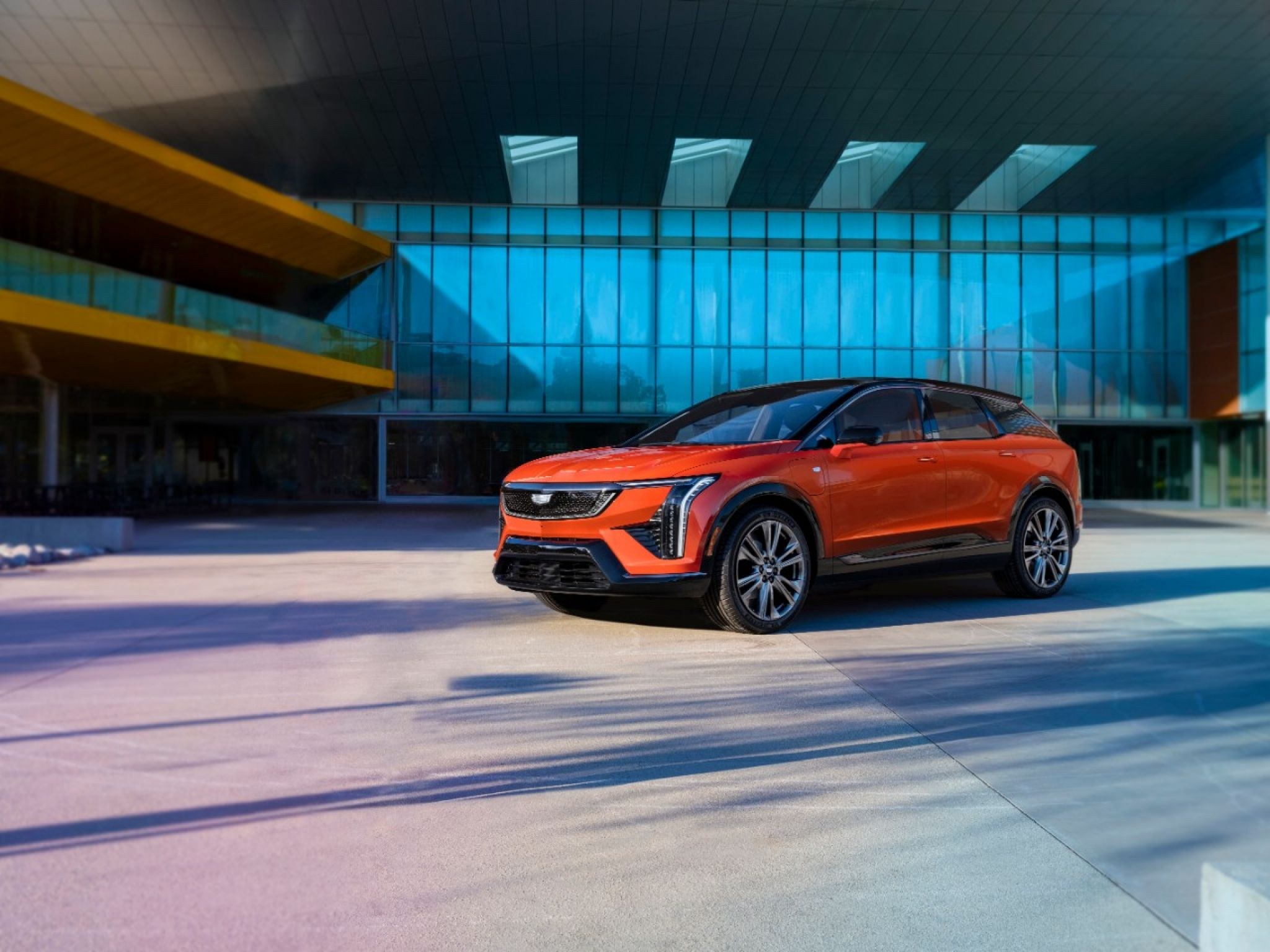 2025 Cadillac Optiq in orange, front three-quarter view outside a glass building, the electric SUV covered by recall 26V612000 over front windows that may not reverse when closed from the key fob