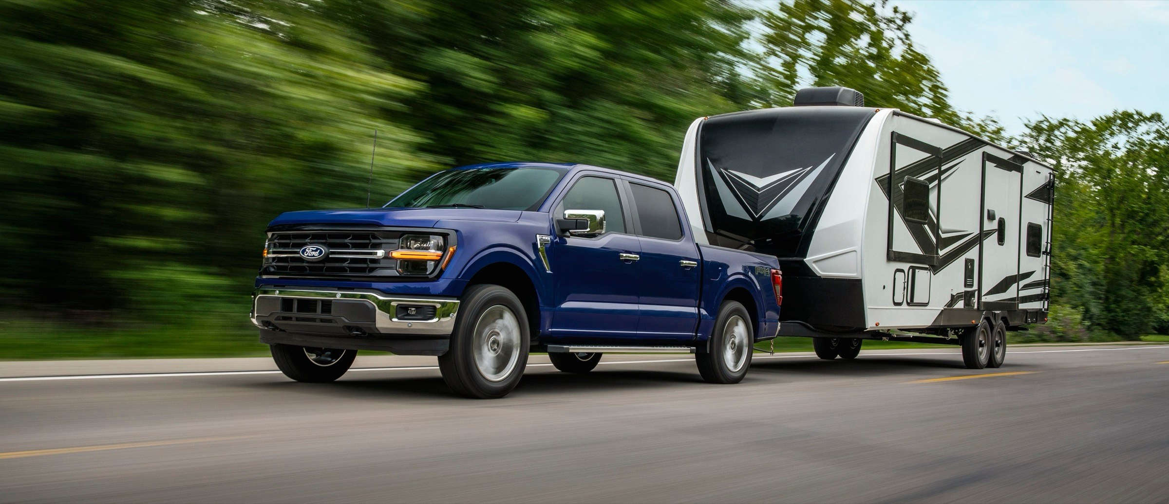 2026 Ford F-150 XLT SuperCrew in blue towing a travel trailer, front three-quarter view, the gas-powered truck generation covered by recall 26V578000 over a front fuel tank strap that may not be seated in the frame