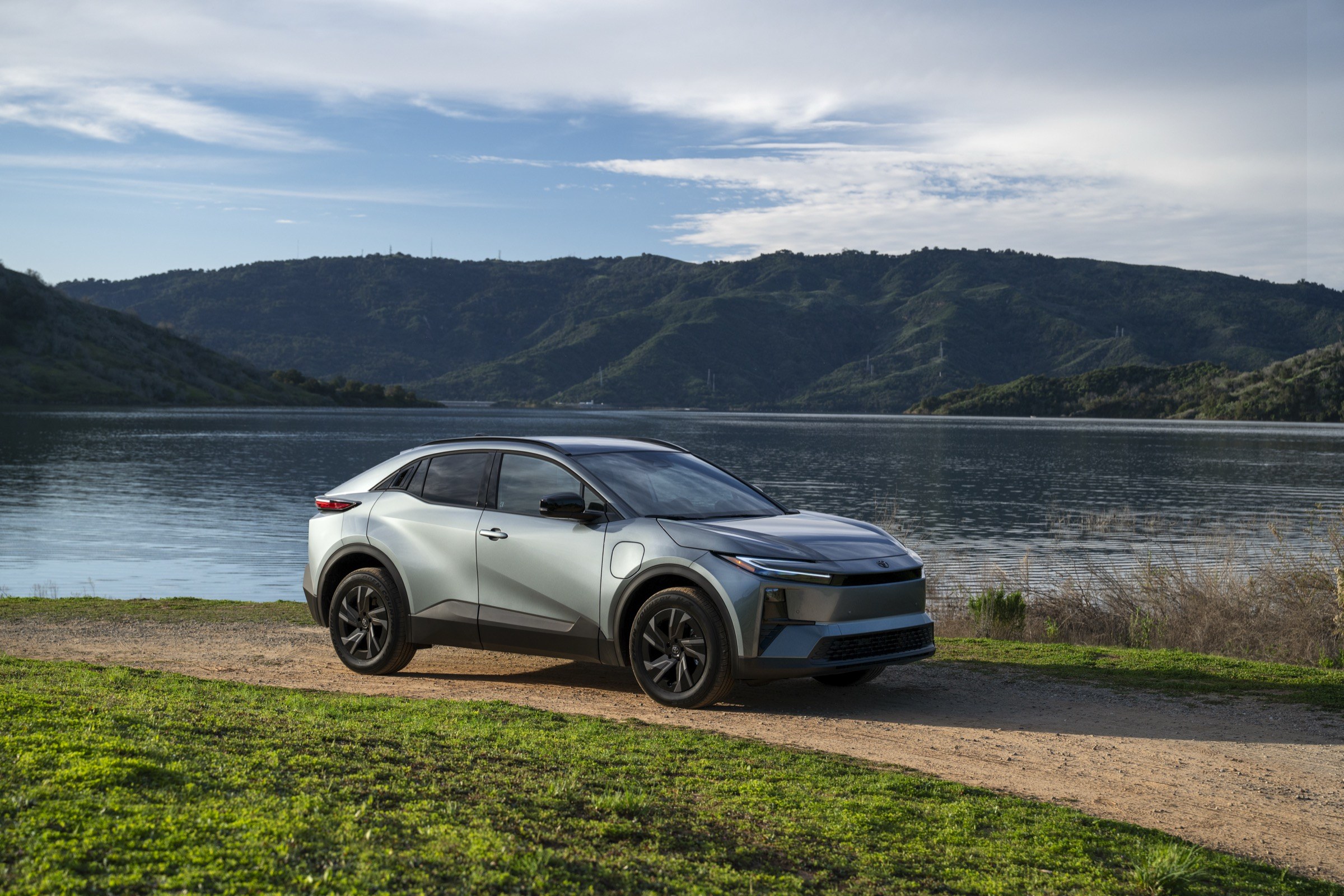 2026 Toyota C-HR SE AWD battery-electric crossover in silver, front three-quarter view beside a lake, the model recalled under campaign 26V570000 because a software error can overcharge the drive battery and cut drive power