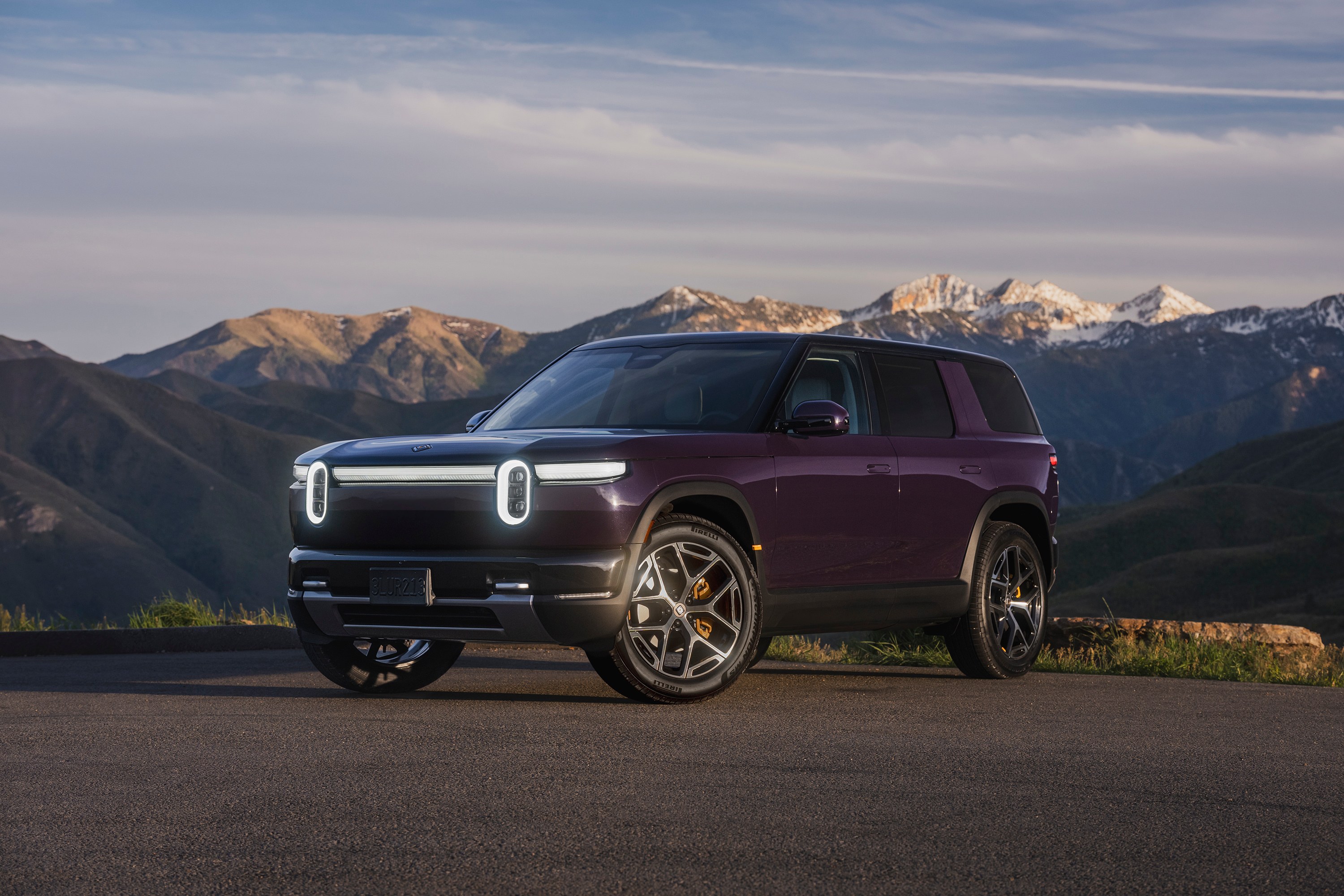 Rivian R2 in dark purple parked on a mountain overlook in Utah, front three-quarter view with headlights on, the model covered by recall 26V625000 over a high-voltage battery fastener