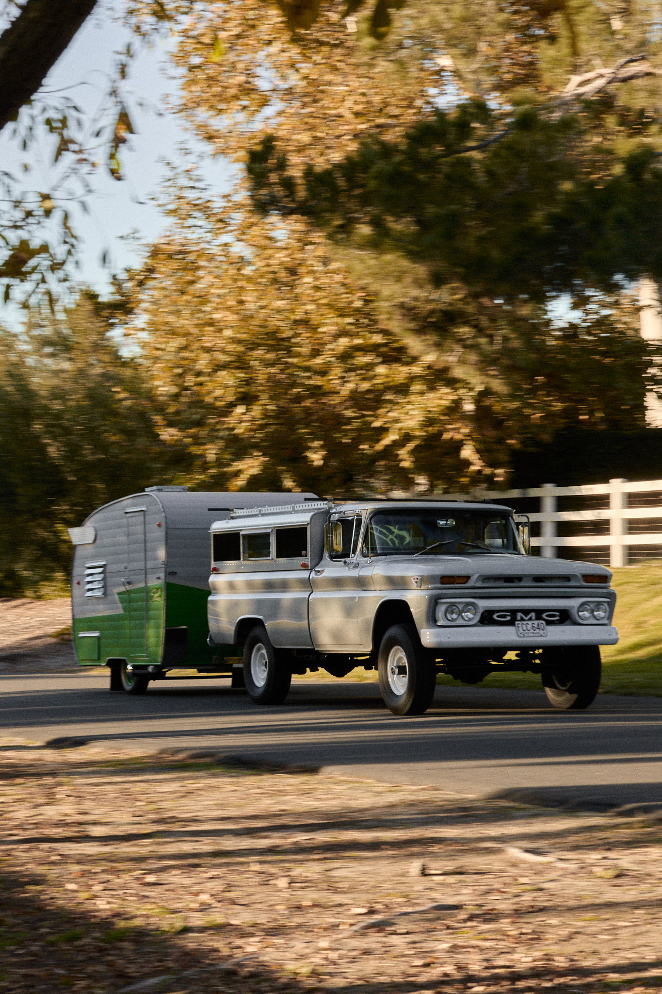 1963 GMC K1000 with custom camper shell and Shasta trailer driving on tree-lined road