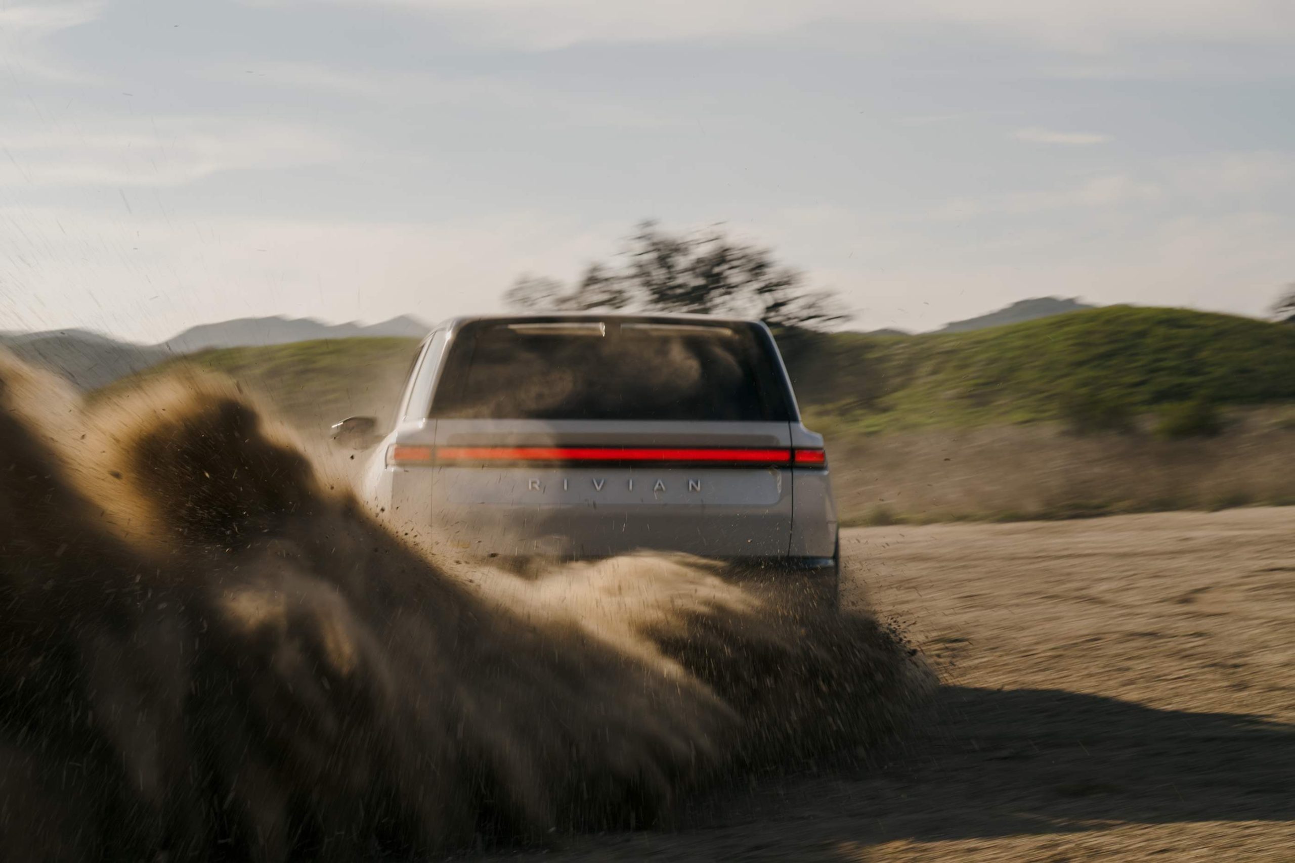 2026 Rivian R2 electric SUV rear view kicking up dirt on a desert trail at golden hour