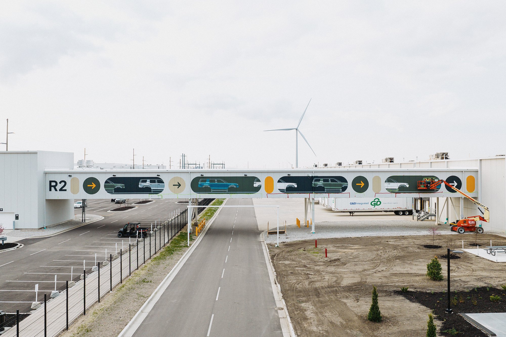 Rivian Normal Illinois plant exterior with R2 mural on pedestrian bridge and wind turbine in background