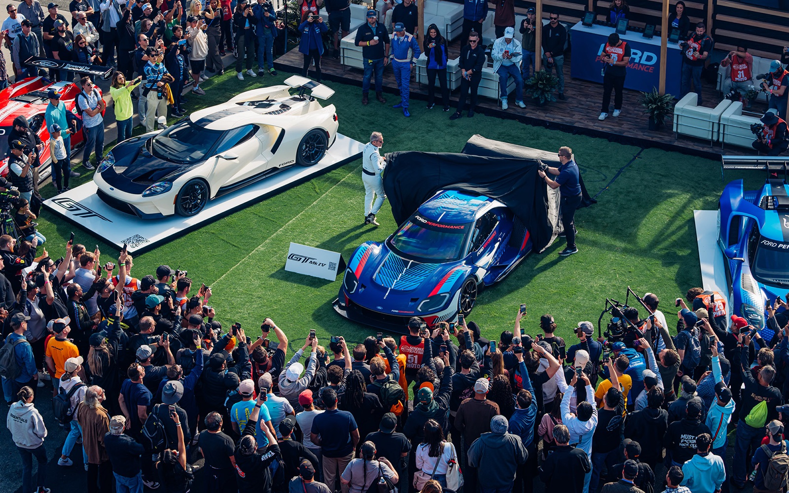 Crowd gathered around a hypercar reveal at Velocity Invitational, photographers and spectators pressed close to a white and a blue supercar on display stands