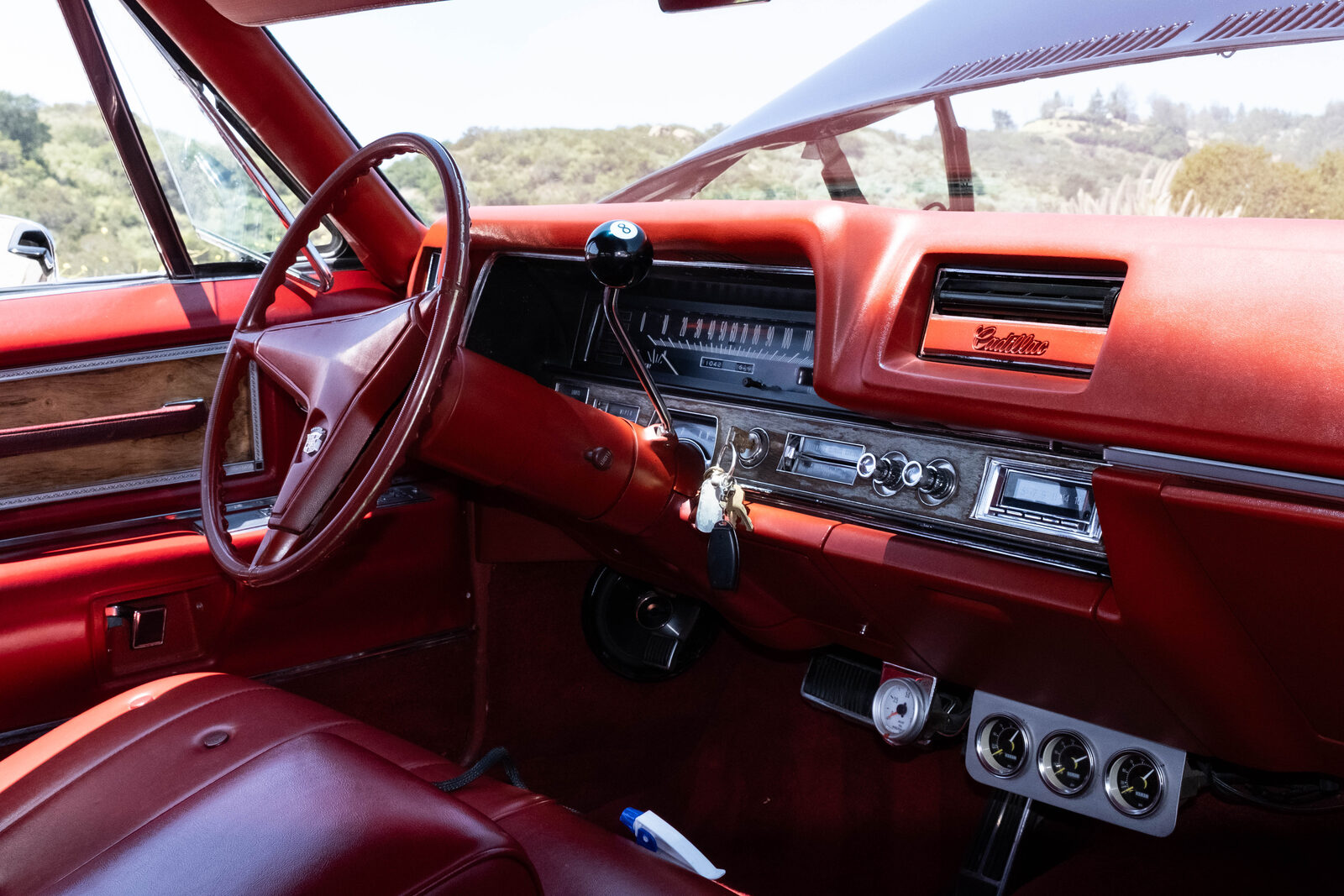 Red vinyl interior of the 1968 Cadillac Coupe DeVille showing the woodgrain steering wheel, retro Bluetooth head unit, and added air-suspension pressure gauges