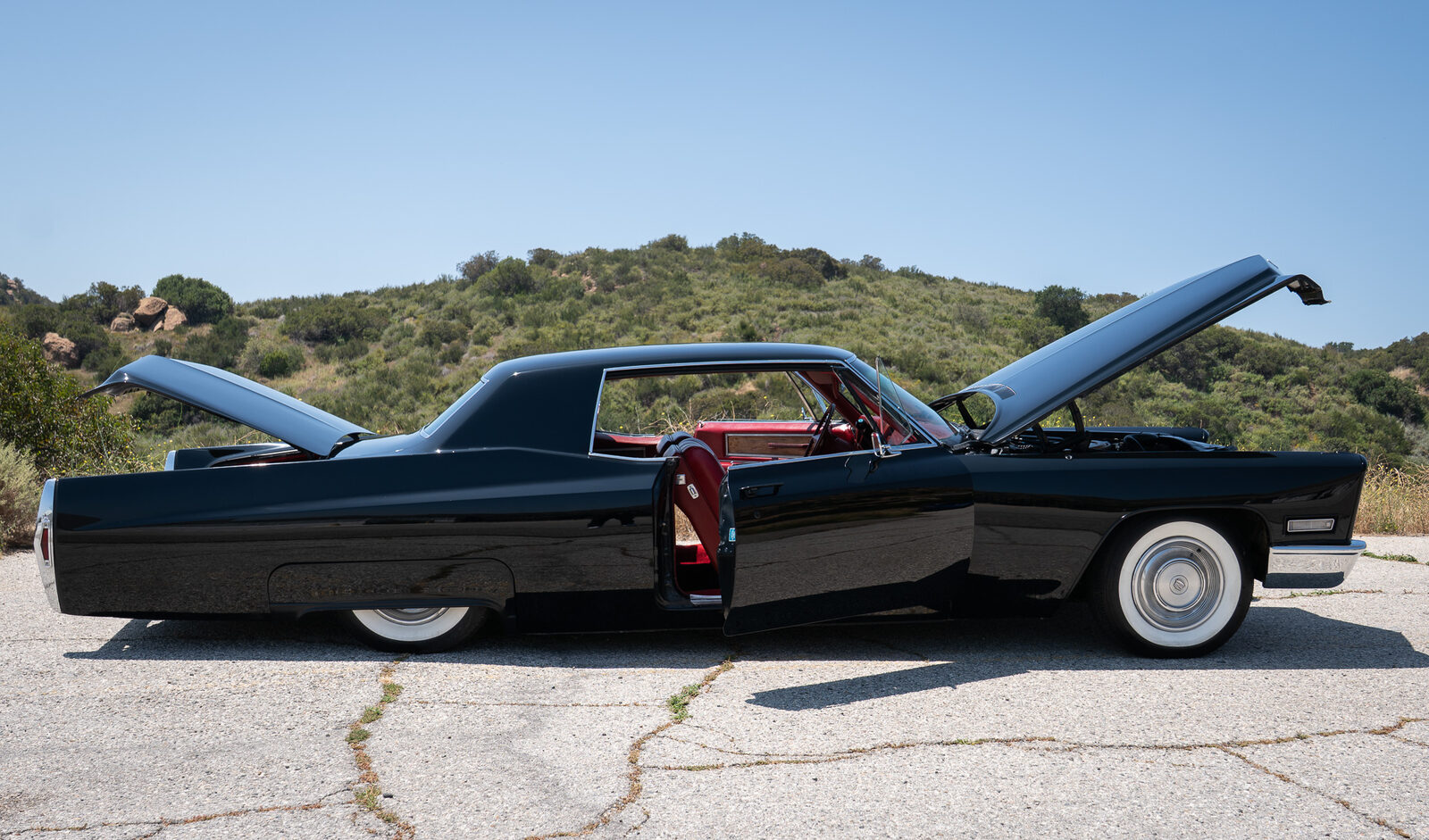 Black 1968 Cadillac Coupe DeVille side profile laid out on its air suspension, doors and hood open showing the red vinyl interior