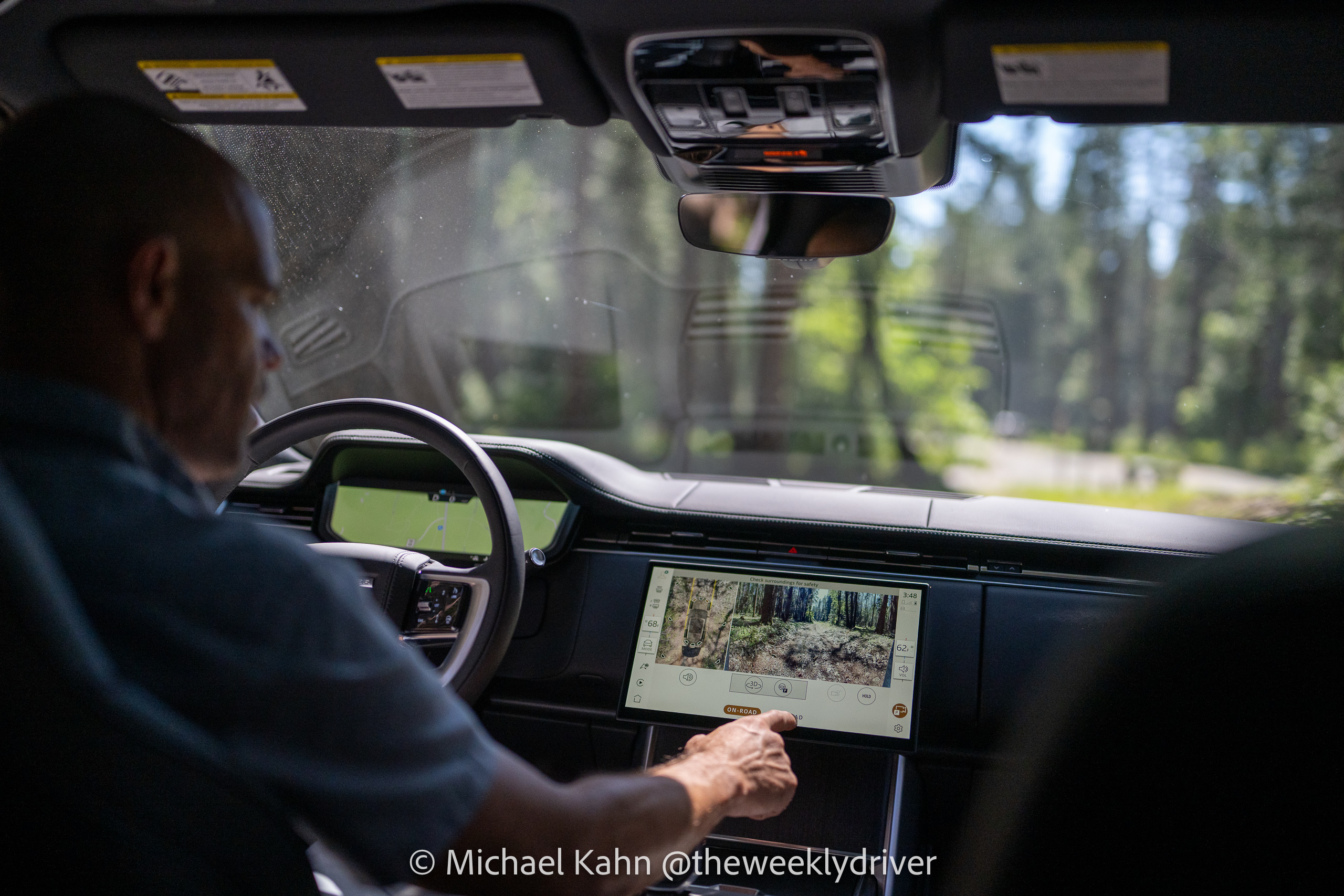 2026 Range Rover SE cabin showing the Pivi Pro 13.1-inch touchscreen with 3D Surround Camera in use on a forest logging road, driver's hand reaching toward the screen