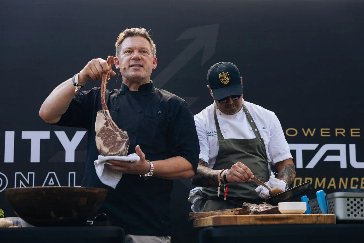 A featured chef holding a tomahawk ribeye steak during a culinary demonstration at Velocity Invitational, with a second chef preparing food beside him