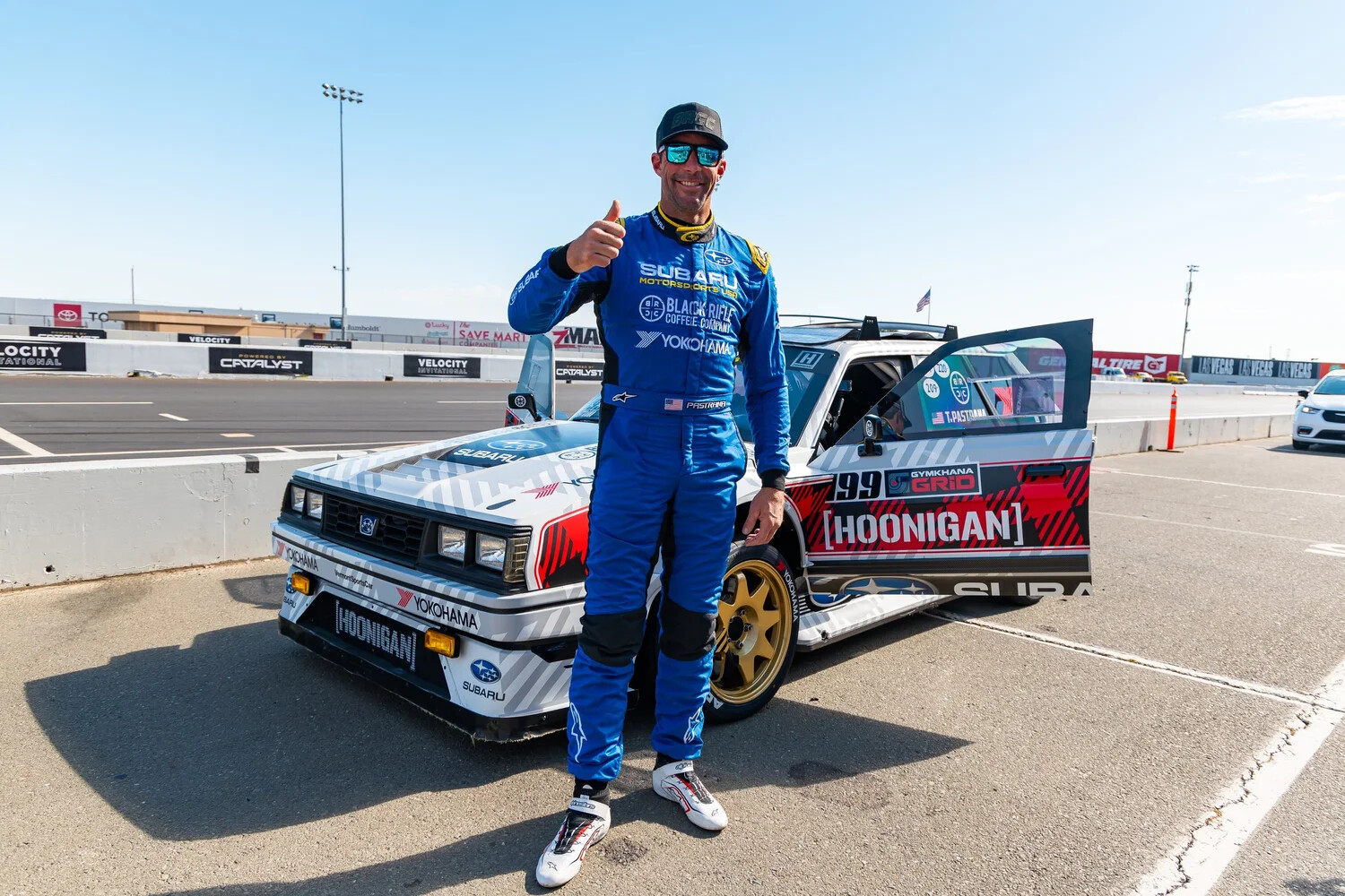 Formula Drift champion Dai Yoshihara in a blue Subaru-livery driver's suit standing in front of his Hoonigan-branded Subaru drift car at Sonoma Raceway