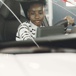 A young driver seated behind the steering wheel of a modern car, viewed through the windshield, the vantage point a cabin-facing driver monitoring camera uses to read the face for drowsiness, distraction, and now heart rate