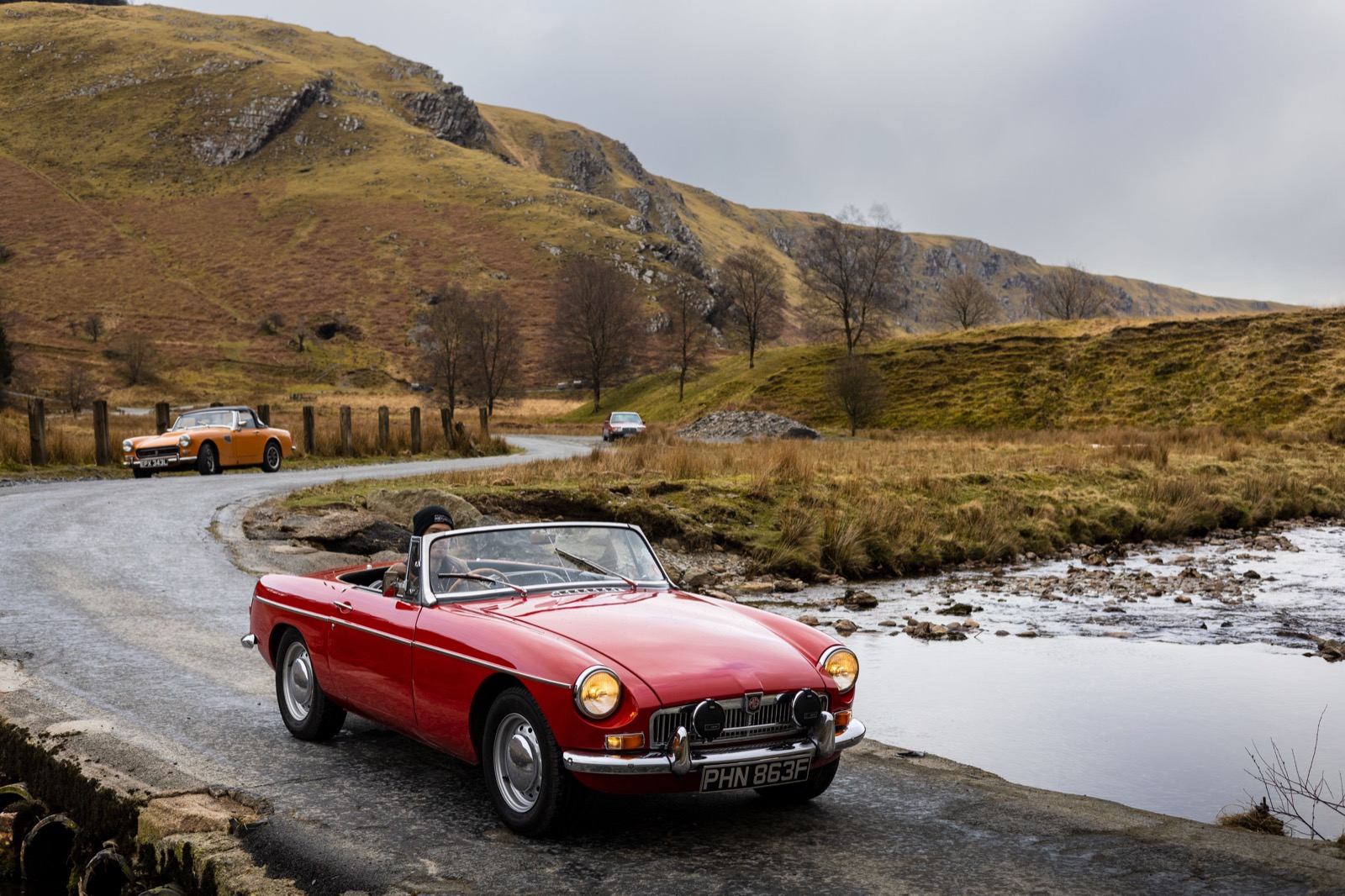 A red MGB leading an orange MG Midget and a classic Mercedes on a mountain road in Wales