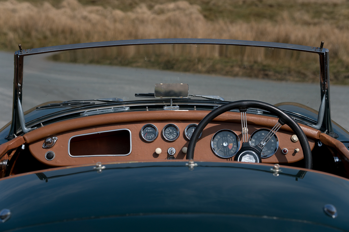 The driver's-eye view over the dashboard of a classic British roadster, showing Smiths gauges and a banjo-spoke steering wheel
