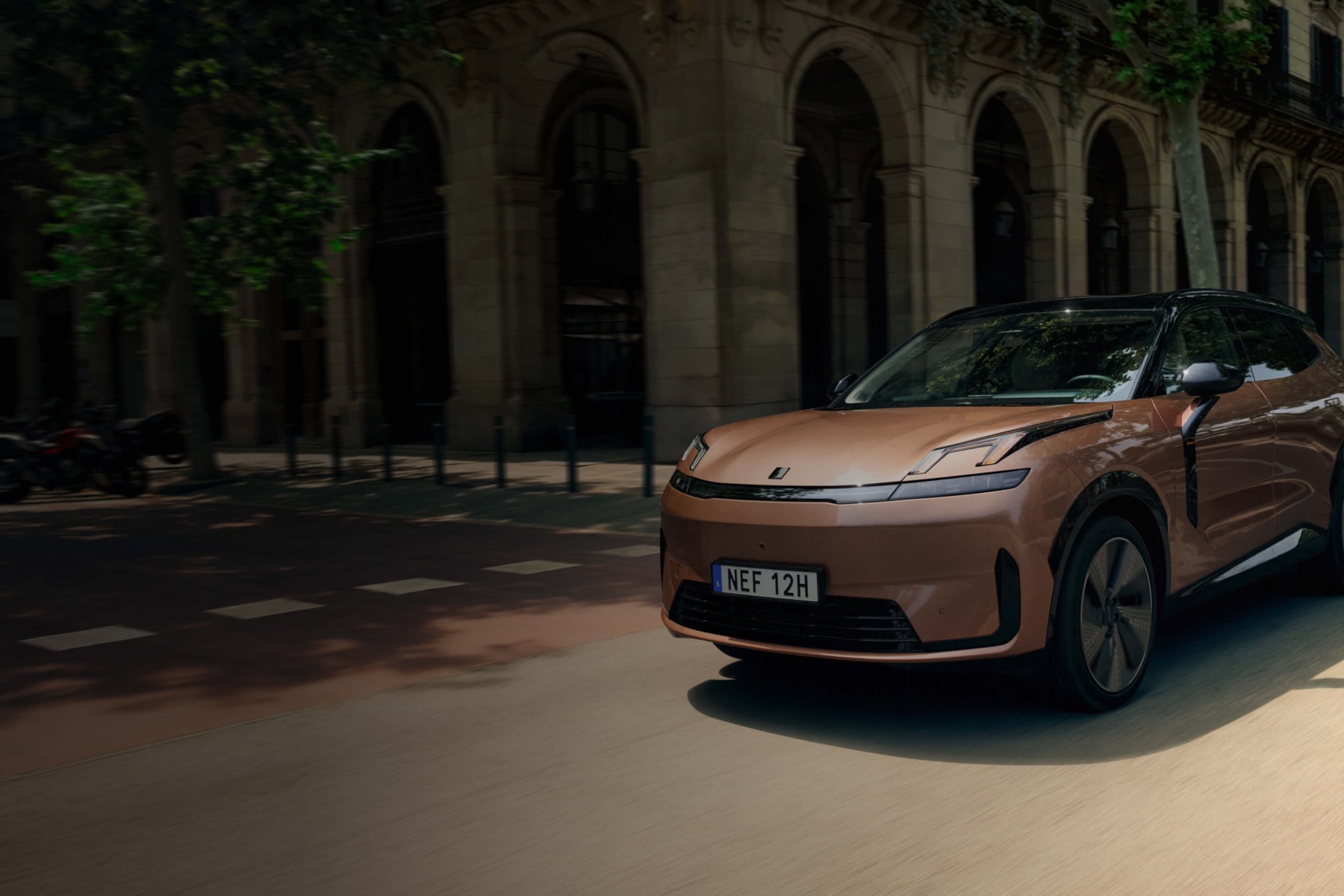 A copper-colored Lynk and Co 08 driving past an arcaded stone building on a European city street