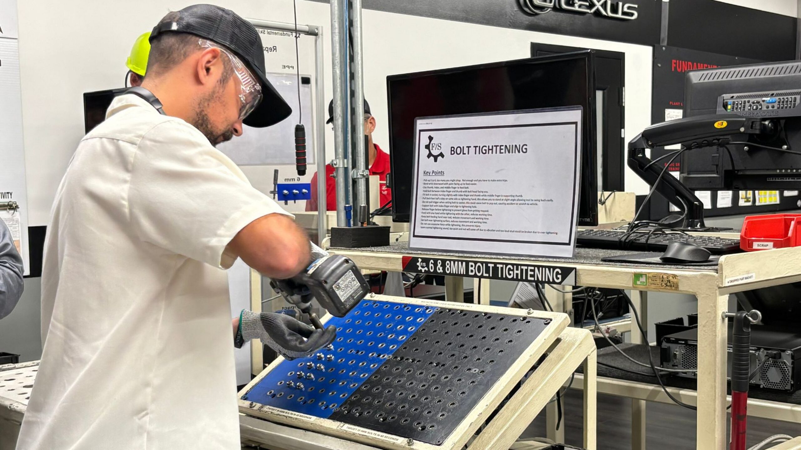 Toyota team member practicing bolt-tightening at a standardized-work training station inside a Toyota plant, with a laminated key-points sheet beside the fixture