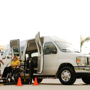 A wheelchair user on a deployed lift boarding a white accessible van while an attendant operates the lift