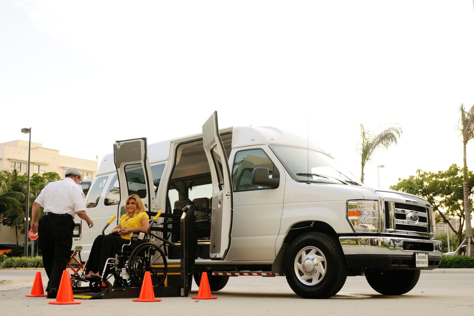 A wheelchair user on a deployed lift boarding a white accessible van while an attendant operates the lift