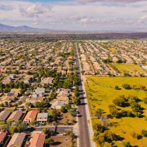 Aerial view of a suburban Arizona arterial road running straight through dense tract housing, with a park, further subdivisions and mountains visible toward the horizon