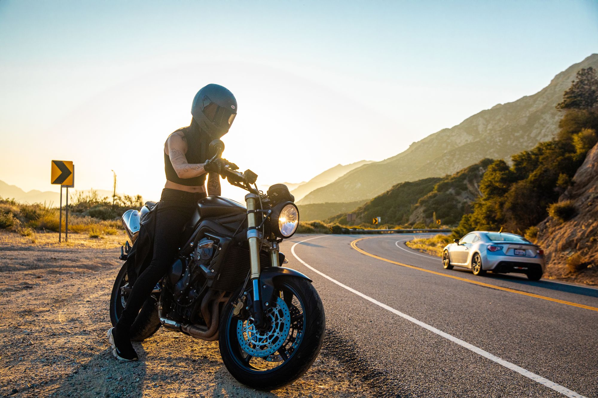 Home 11 Motorcyclist stopped on the shoulder of a winding canyon road at sunset as a car passes on the curve
