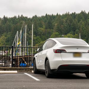 White 2023 Tesla Model Y parked in a waterfront lot in Gig Harbor, Washington, with sailboat masts and a forested hillside behind it