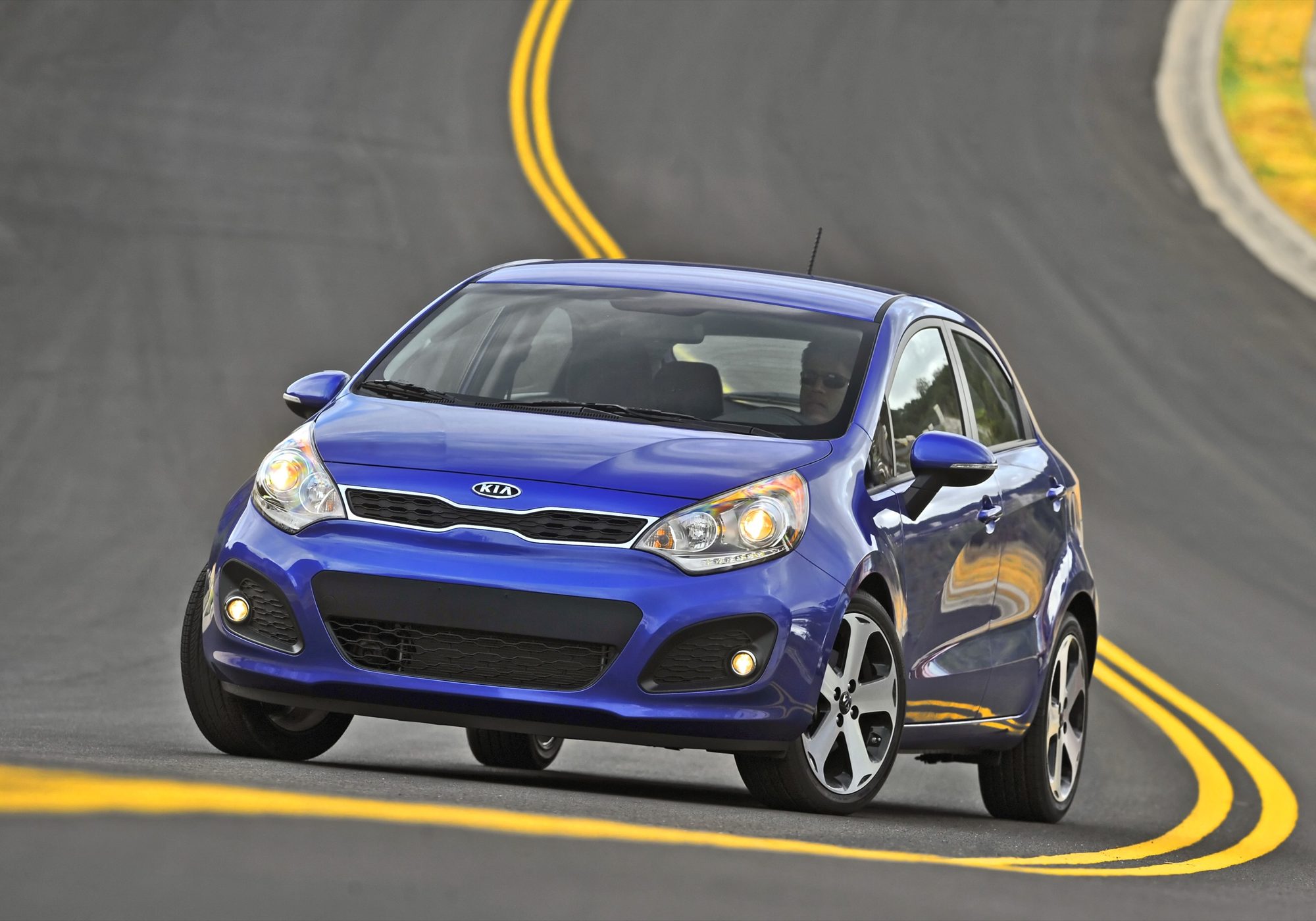 Blue 2012 Kia Rio SX 5-door cornering on a two-lane road