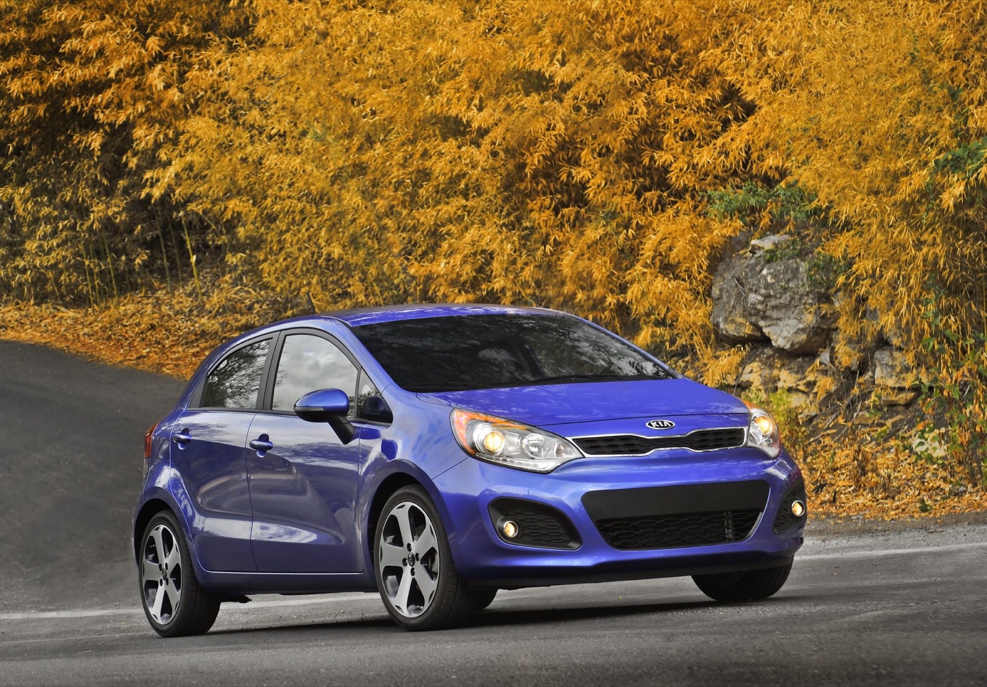 Blue 2012 Kia Rio SX 5-door hatchback driving on a road lined with autumn foliage