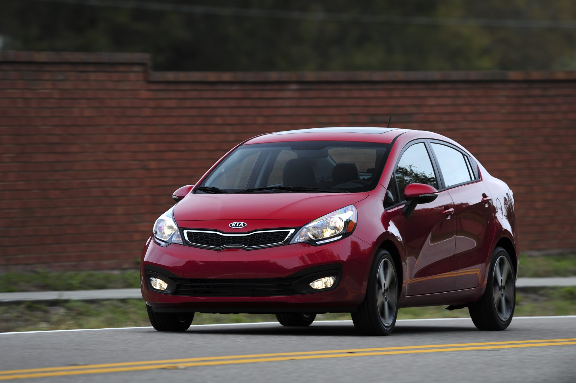 A red 2012 Kia Rio SX sedan driving on a curving road