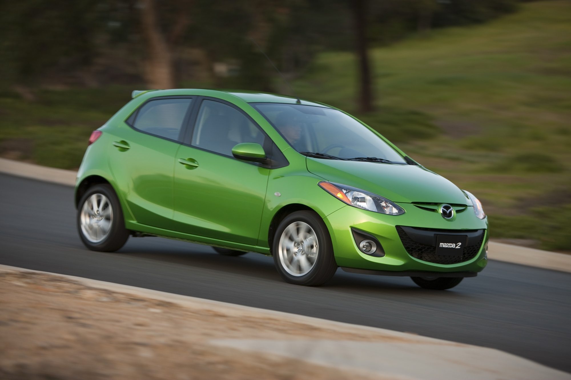 A green 2012 Mazda2 driving on a curving road
