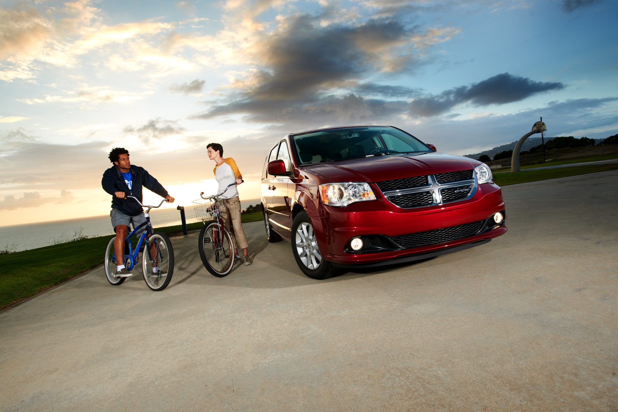 Front three-quarter view of a red 2013 Dodge Grand Caravan Crew parked by the ocean with two cyclists