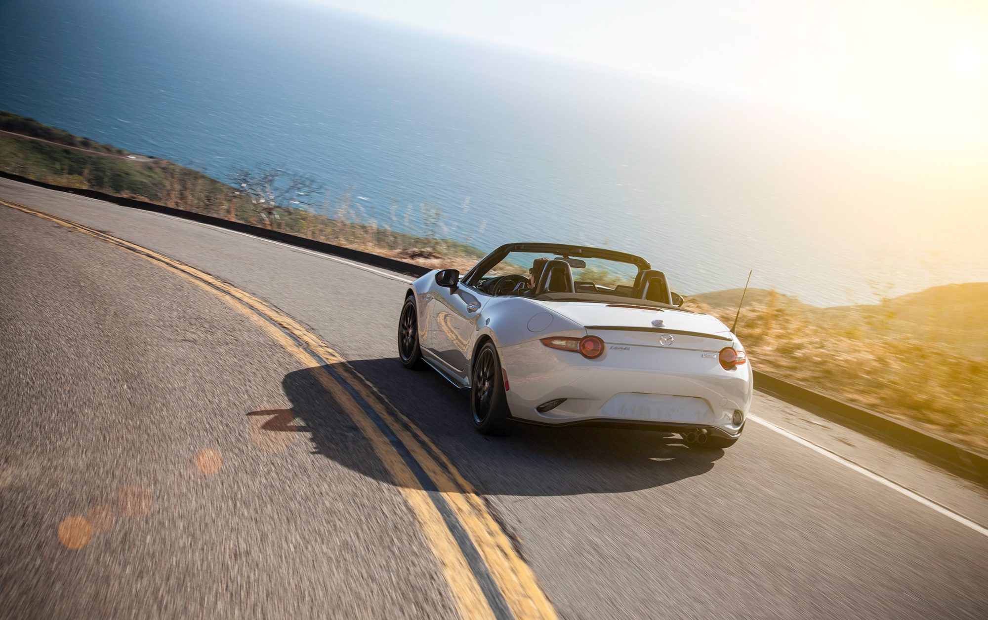 White 2016 Mazda MX-5 Miata Club with the top down on a coastal road at sunset