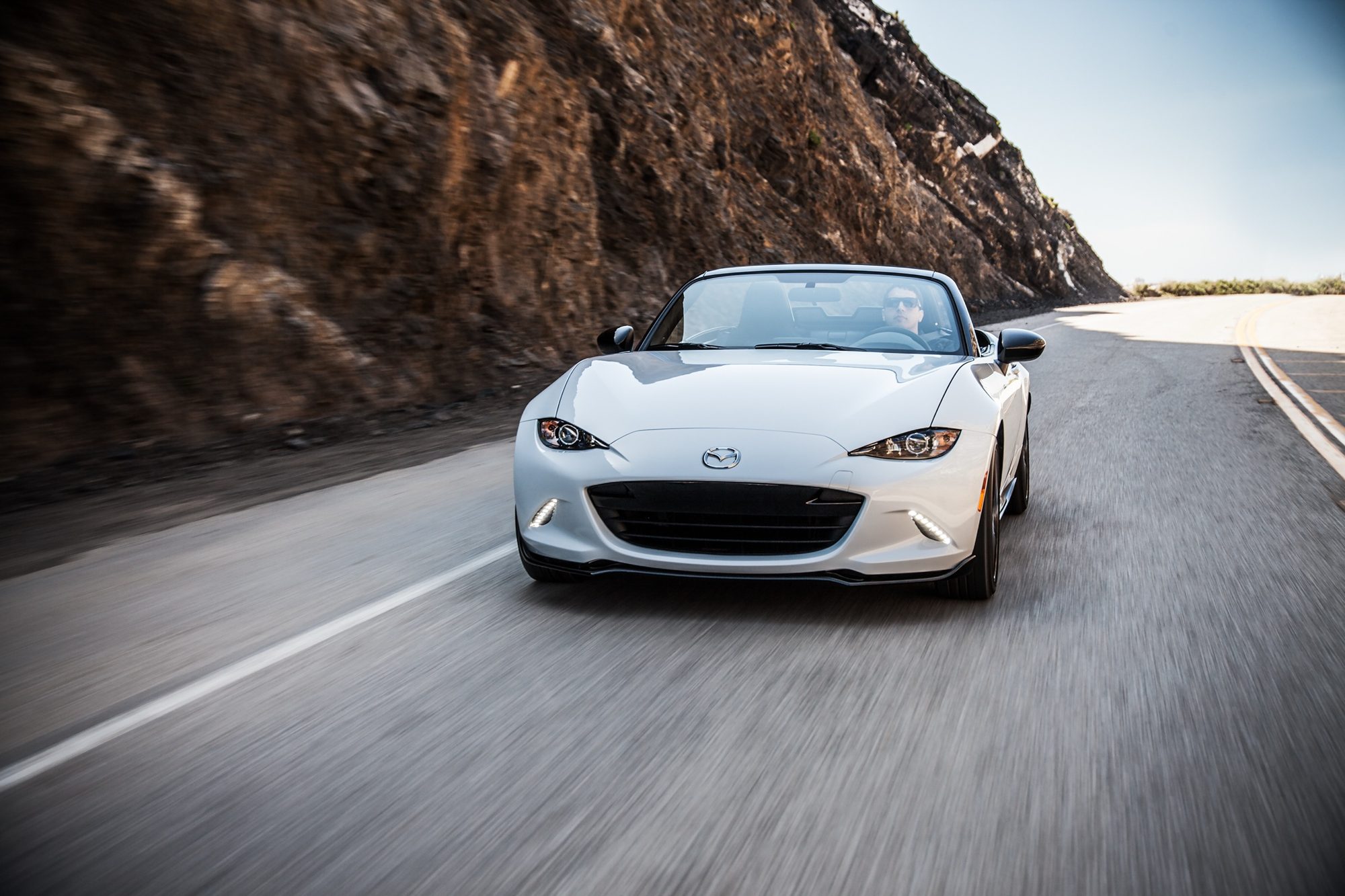 White 2016 Mazda MX-5 Miata Club driving on a winding coastal road with the top down