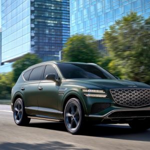 Dark green Genesis GV80 Hybrid driving past glass office towers, front three-quarter view