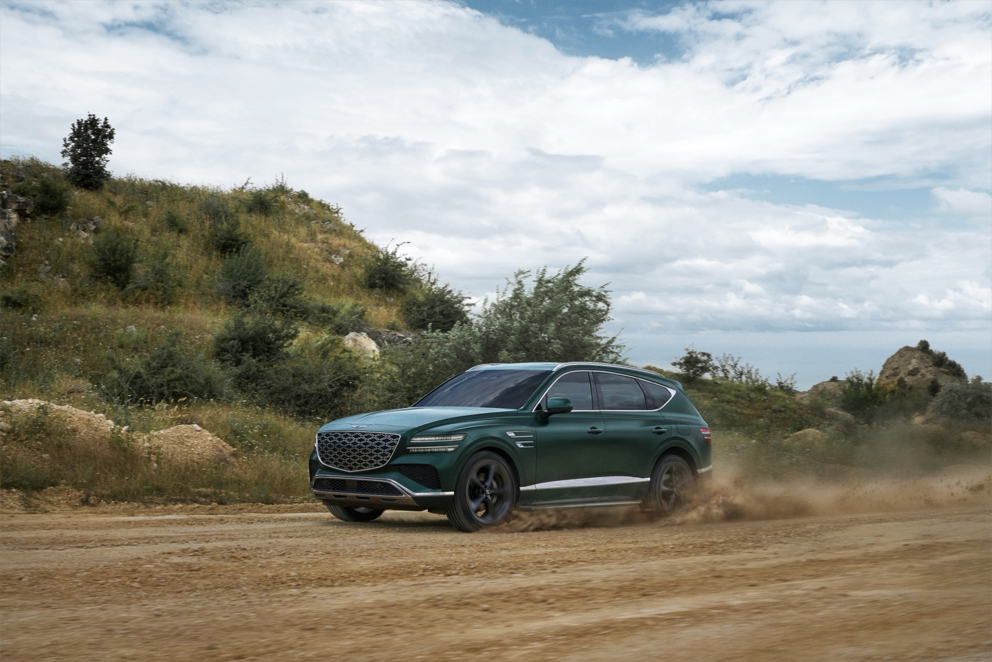 Dark green Genesis GV80 Hybrid kicking up dust on a gravel road, side profile