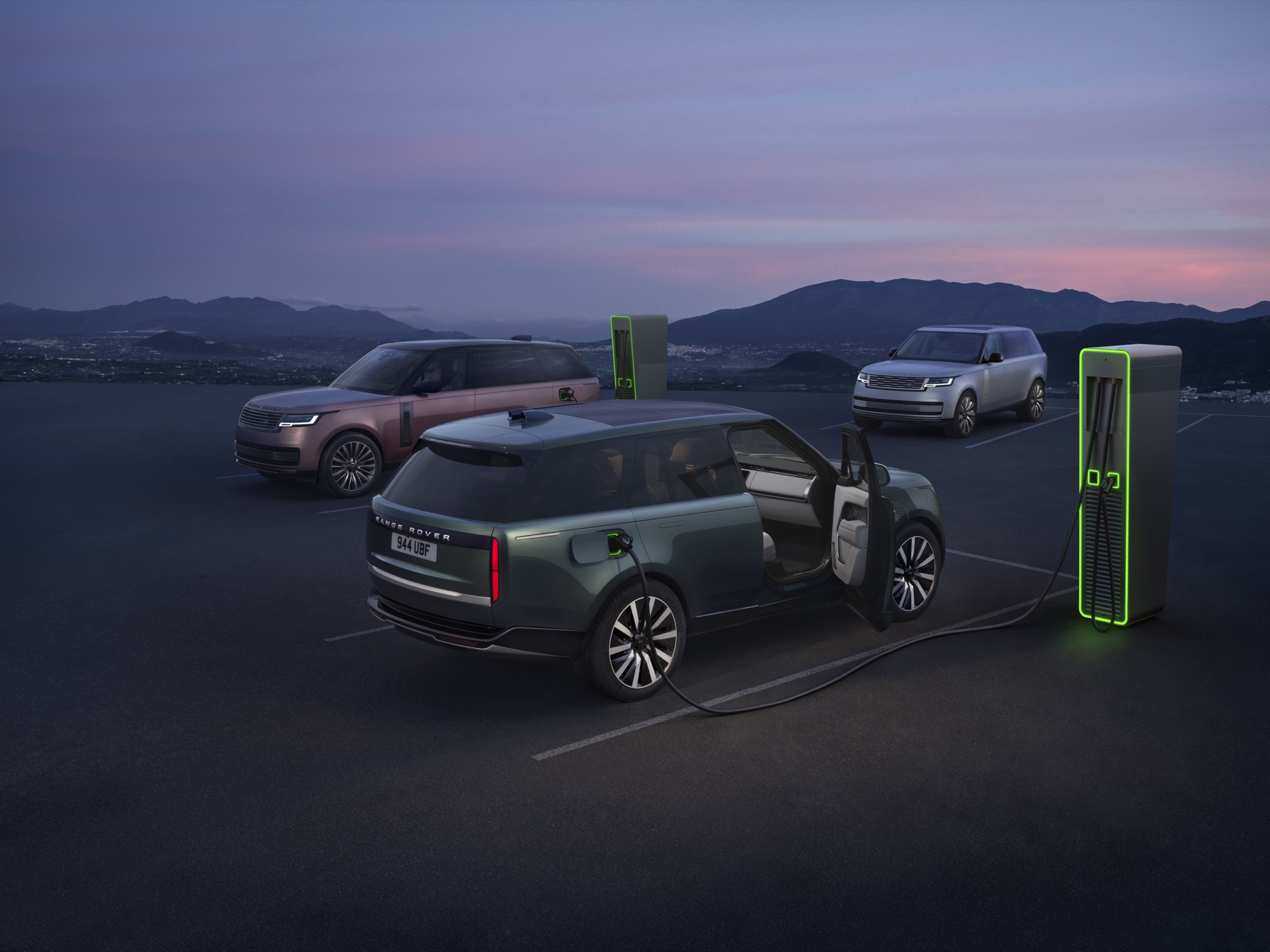 Three Range Rover Electric models at charging posts at dusk, the nearest with its driver door open and a cable plugged in