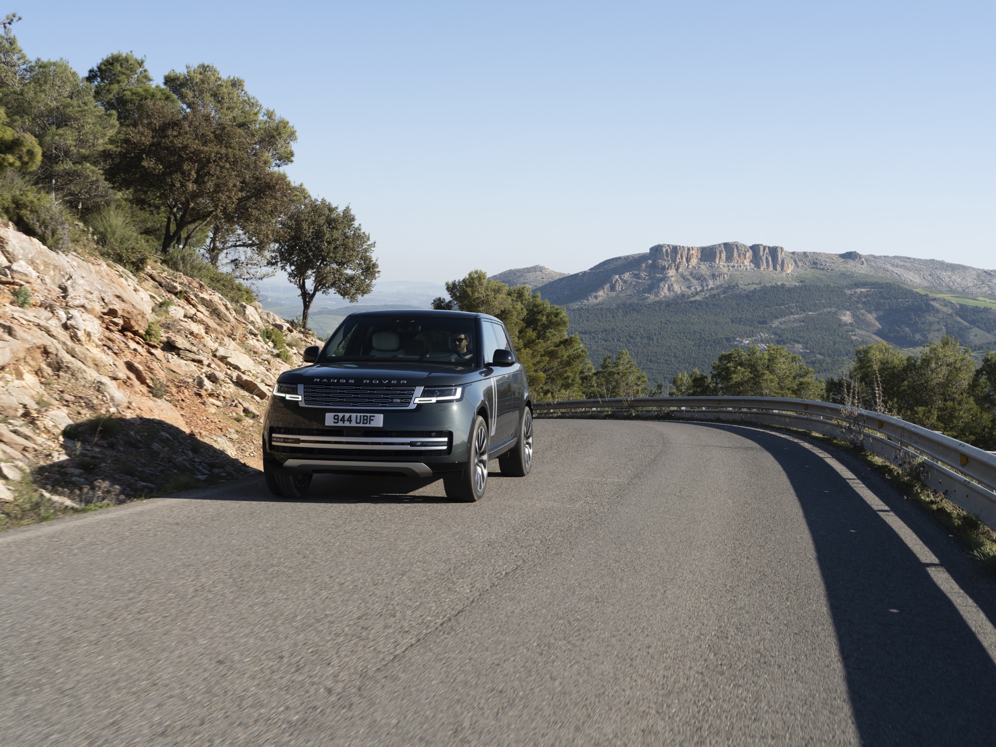 Range Rover Electric Starts at $138,000, the Same Price as the V8 SE 13 Dark green Range Rover Electric driving on a mountain road with a rock outcrop behind