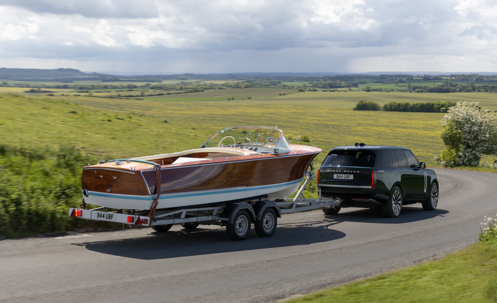 Dark green Range Rover Electric towing a wooden speedboat on a trailer along a country road