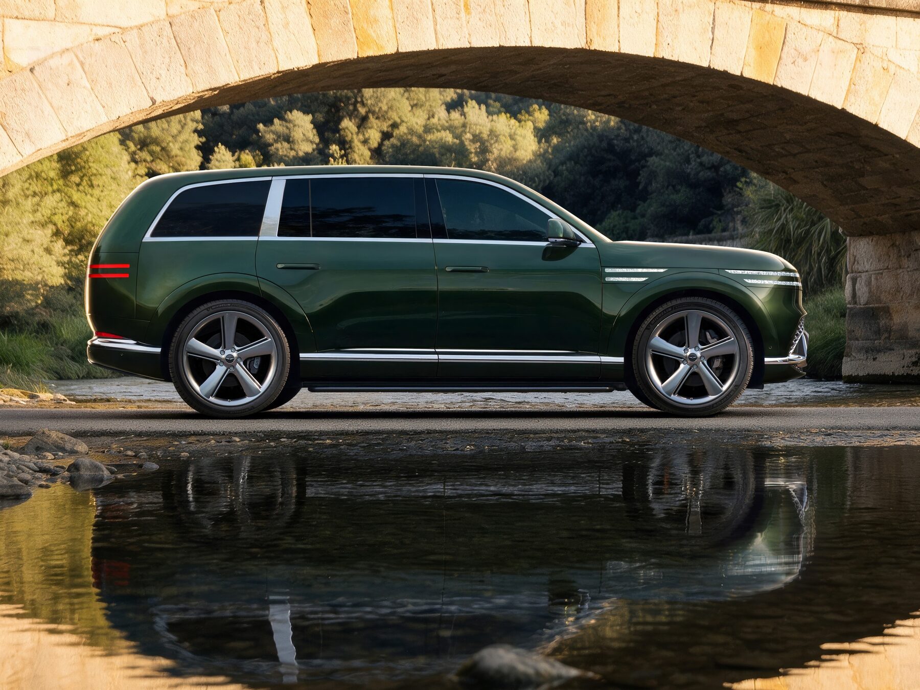 Dark green Genesis GV90 in side profile beneath a stone bridge, reflected in water
