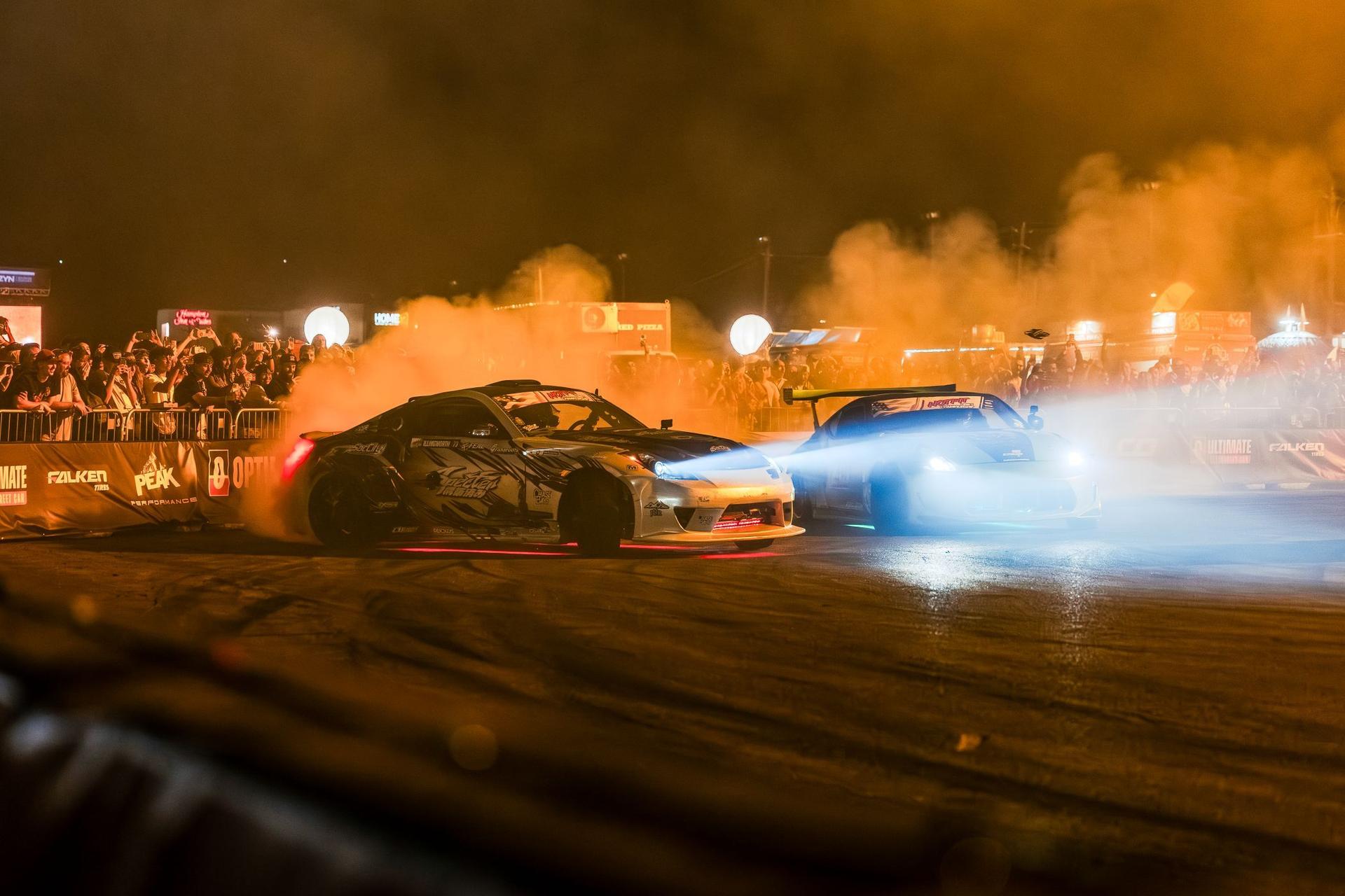 Two drift cars slide side by side in tire smoke in front of a crowd at SEMA Fest