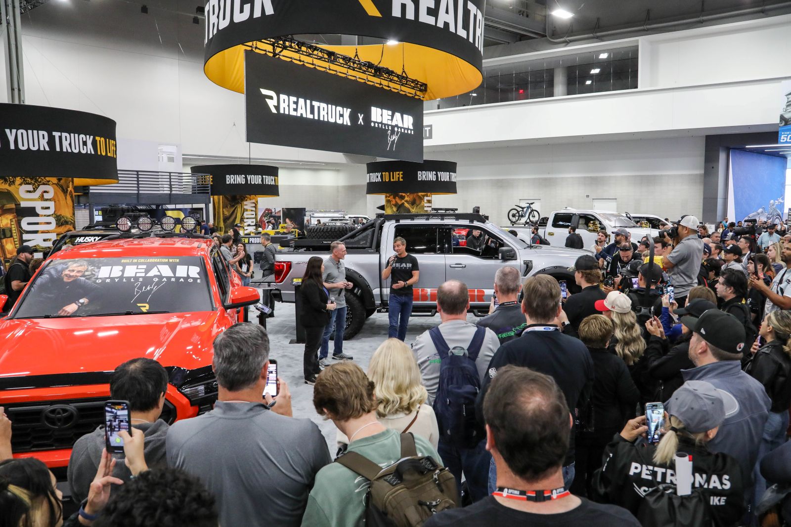 A crowd photographs two modified pickup trucks at the RealTruck booth during a SEMA Show presentation